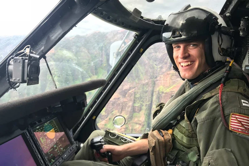 A smiling man in a white pilot's helmet and olive green flight suit, wearing an American flag patch, pilots a helicopter. He grins from the cockpit, holding a control stick, with lush green, rocky cliffs visible beyond the window.