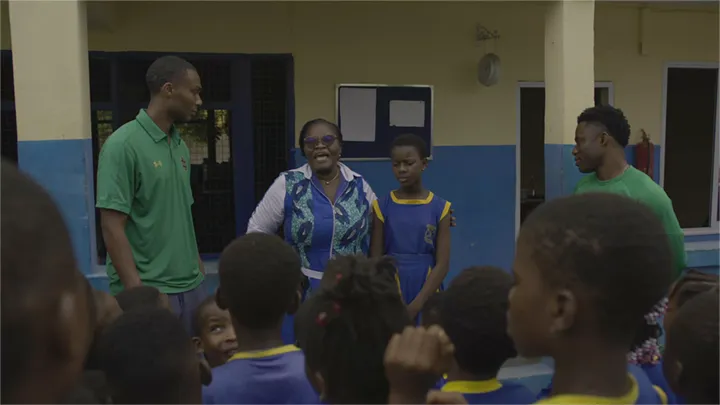 A group of young students in blue and yellow uniforms face a woman in a patterned top and three men in green shirts and a red scrub top in a schoolyard setting.