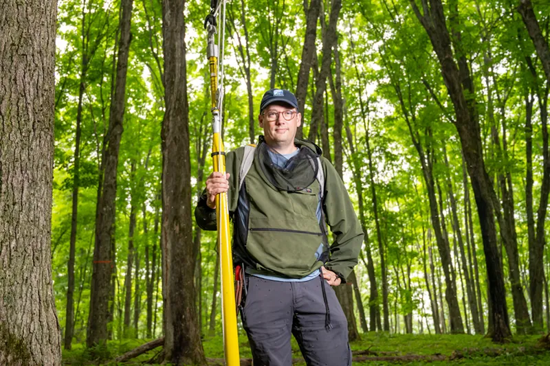 Nate Swenson stands in a lush green forest, holding a tall yellow pole saw. He wears a green jacket, grey pants, glasses, and a baseball cap with bug netting around his neck. Mosquitoes are visible swarming on his clothing. Sunlight filters through the dense canopy of trees behind him.