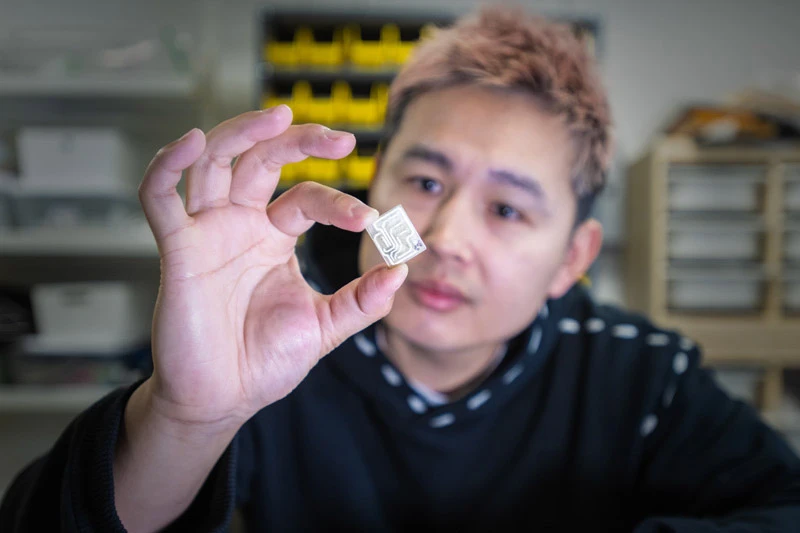 A man with spiked reddish-brown hair holds a small, clear square chip with internal serpentine channels, examining it intently. He wears a dark hoodie in a lab setting.