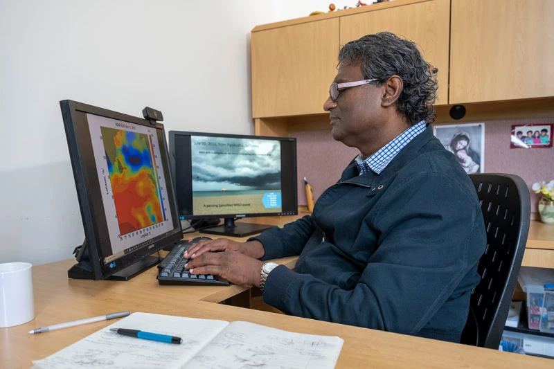 A man with grey curly hair and pink glasses types on a keyboard at a desk, looking at two monitors. The left screen shows a colorful scientific map. The right shows a dark, cloudy beach scene from Sri Lanka and text about an MJO event.