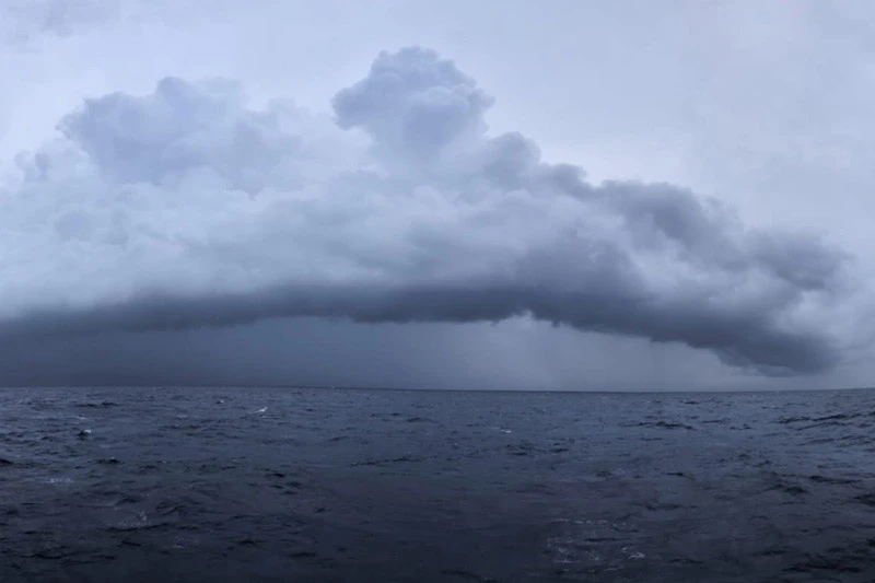 A wide expanse of dark, choppy ocean water under a heavy, grey storm cloud with visible rain falling in the distance.