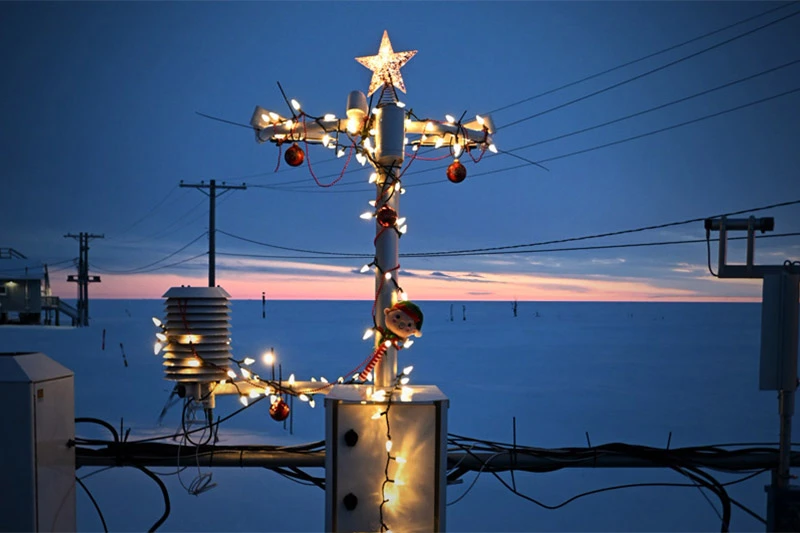 A white pole with a lit star, string lights, red ornaments, and an elf figure, along with a lit weather station, stands against a twilight sky. The blue sky fades to pink above a vast white landscape dotted with distant utility poles and wires.