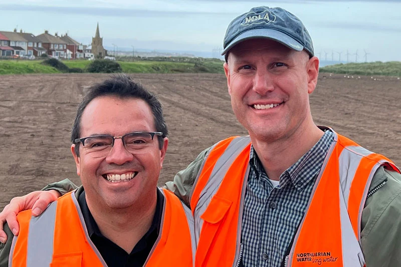Two smiling men in orange safety vests stand outdoors. The man in a NOLA cap has his arm around the other. Behind them are a dirt field, houses, a church steeple, and distant wind turbines.