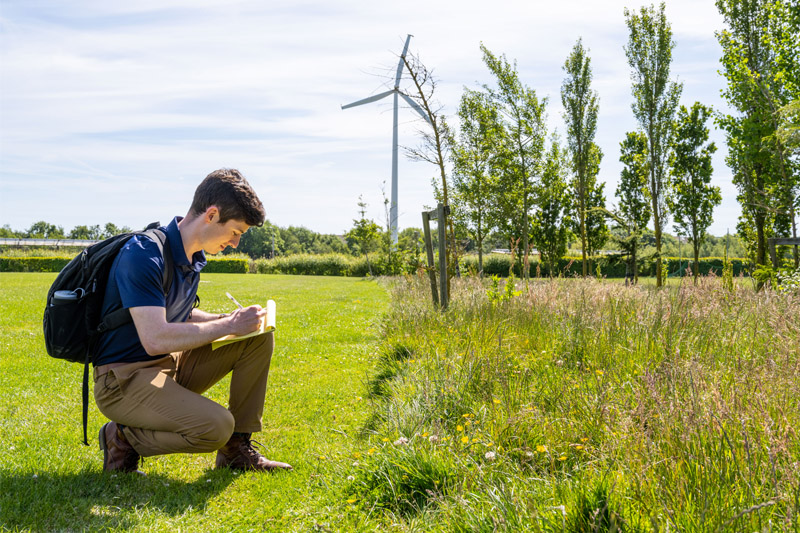 A young man wearing a blue polo shirt and tan pants kneels in a grassy field, writing on a yellow notepad. He wears a black backpack and works in front of a tall white wind turbine and a row of green trees under a bright blue sky.