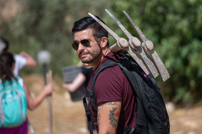 A bearded man wearing sunglasses and a backpack looks over his shoulder outdoors, with three large digging tools or pickaxes attached to his pack.