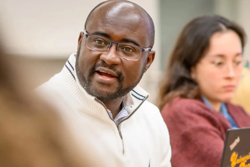 A bald, dark-skinned man with a beard and blue glasses, wearing a white quarter-zip with navy collar stripes, speaks with an engaged expression. A woman in a maroon sweater uses a laptop in the blurred background.