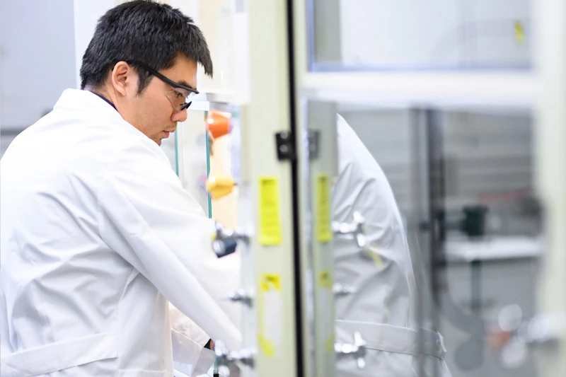A man in a white lab coat and black glasses intently works inside a laboratory fume hood. Orange and yellow equipment are visible within.