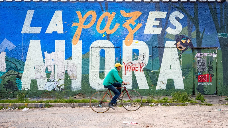A man in a yellow cap and turquoise shirt rides a red bicycle on a street. Behind him, a vibrant mural reads &ldquo;LA PAZ ES AHORA&rdquo; in white and orange over a blue and green painted landscape, with a graffitied door.