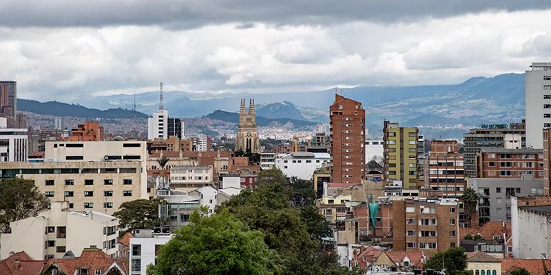 An aerial view of Bogotá, Colombia, featuring the Church of San Francisco with its twin spires, surrounded by residential and commercial buildings, against a backdrop of mountains and a cloudy sky.