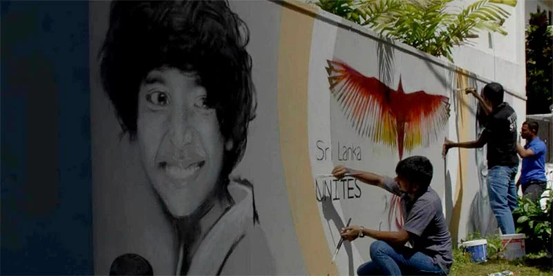 Three men paint a mural. One crouches beside a vibrant red and yellow bird. Another man paints a vertical stripe, and a third watches. Left of the mural is a large, grayscale portrait of a young person with dark hair and wide eyes.
