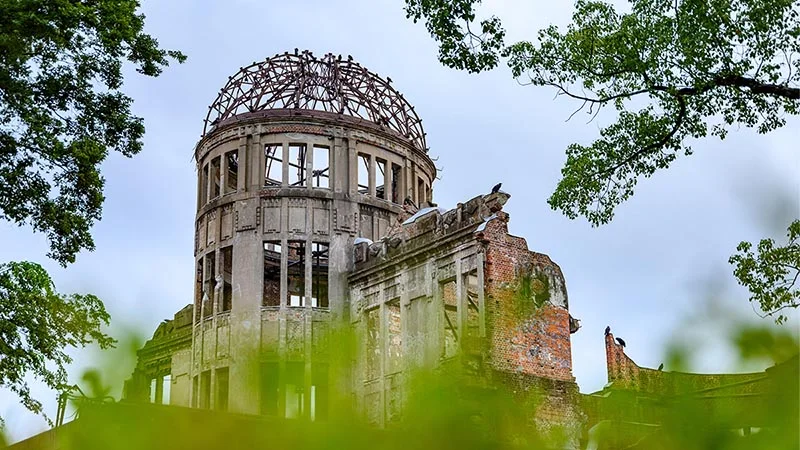 The Hiroshima Peace Memorial, also known as the Genbaku Dome, a skeletal structure with a bare metal dome and crumbling walls, framed by green leaves against a pale sky.
