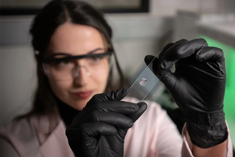 A researcher in a lab coat, safety glasses, and black gloves holds up a glass microscope slide containing a small biological sample.