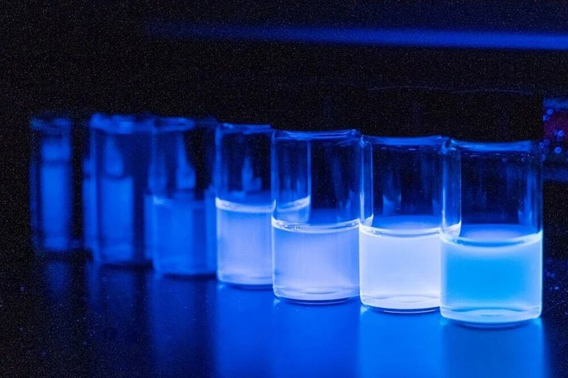 A row of small glass vials containing translucent liquid glows with a bright blue luminescence under UV light in a dark laboratory setting.