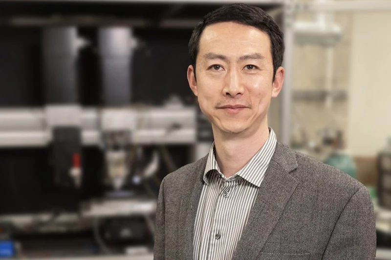 An Asian man with a gentle smile, wearing a striped shirt and gray jacket, standing in a blurred lab with scientific equipment.