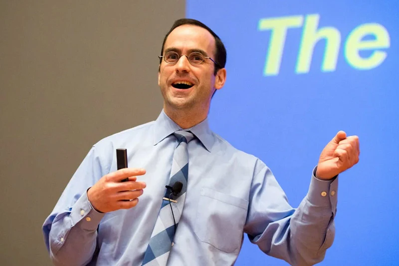 A man with glasses and a blue striped tie smiles enthusiastically while holding a remote clicker during a presentation in front of a blue screen.