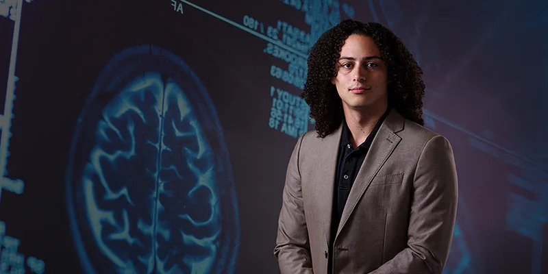 A young man with curly dark hair and a tan suit jacket looks directly at the viewer, standing before a dark blue background displaying a glowing brain scan and digital medical data.