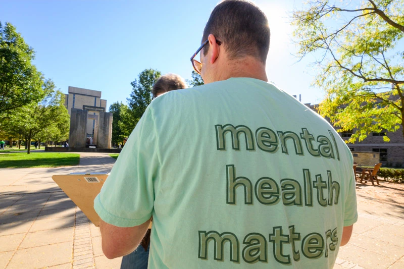 Rear view of a person in a light green &ldquo;mental health matters&rdquo; t-shirt, holding a clipboard on a sunny day at Notre Dame. The Word of Life mural and Hesburgh Library are in the background.