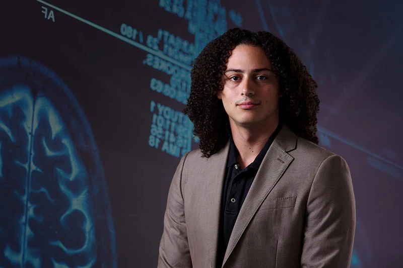 A young man with curly dark hair and a tan suit jacket looks directly at the viewer, standing before a dark blue background displaying a glowing brain scan and digital medical data.