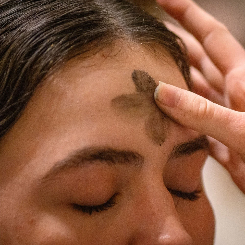 A hand applies dark grey ashes to a person's forehead, forming a cross. The person's eyes are closed.