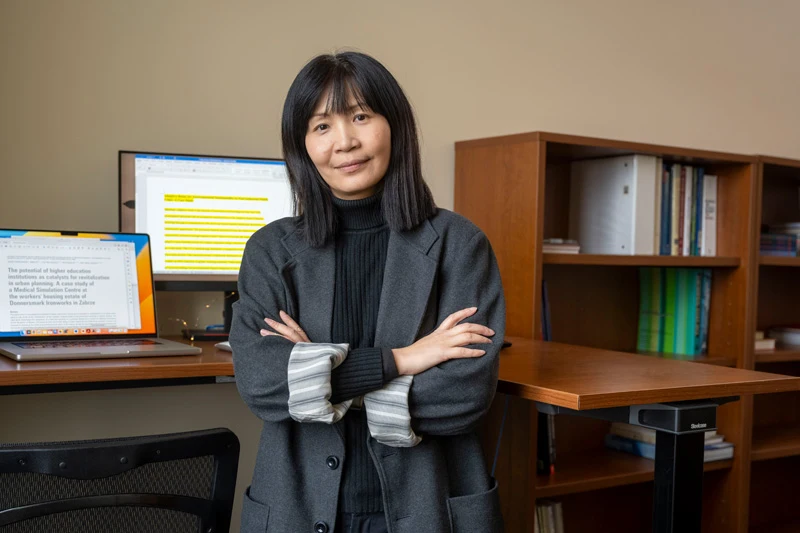 A woman with long dark hair stands with crossed arms in an office, with two computer screens and a bookshelf behind her.