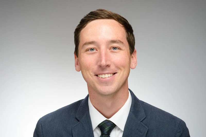 Headshot of a smiling man with short brown hair and blue eyes, wearing a dark blue suit and plaid tie.