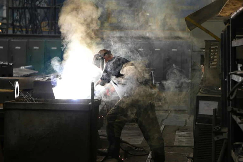 A worker wearing protective gear welds in a factory, generating bright sparks and smoke.