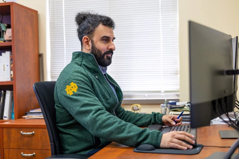 A man with a beard and hair in a bun wears a green Notre Dame pullover while working at a computer desk in an office. 