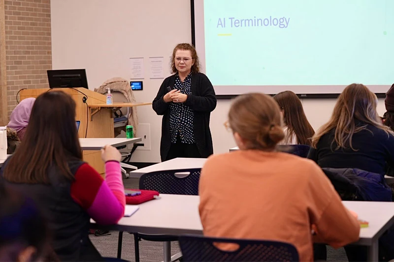 An instructor stands at the front of a classroom lecturing near a projection screen that displays the heading AI Terminology.  