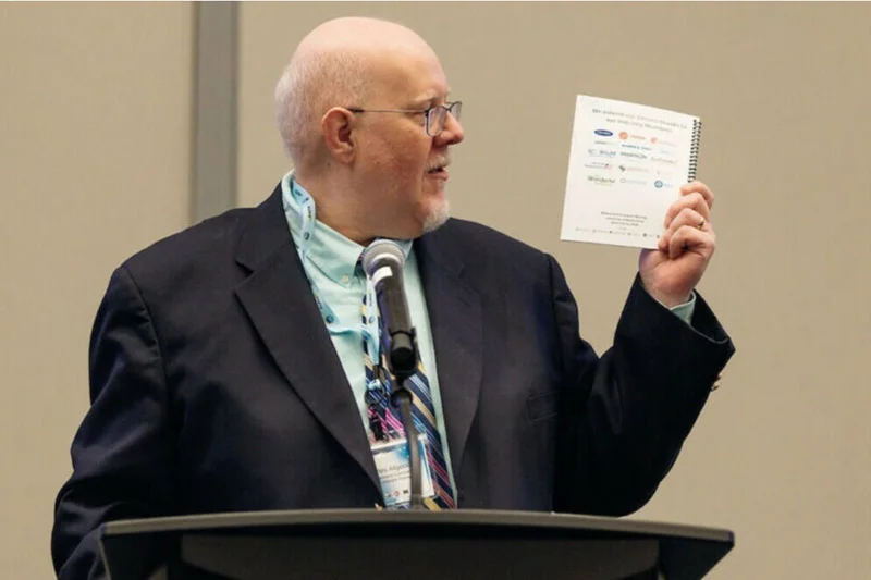 TA bald man in glasses, dark suit, and light blue shirt speaks at a microphone, holding up a white spiral booklet of logos.