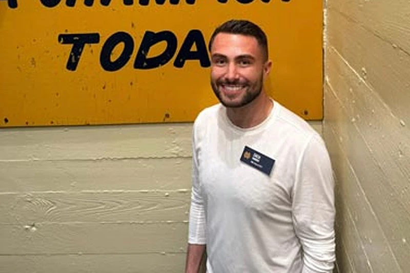 A smiling man in a white long-sleeve shirt stands in a stairwell next to a yellow sign that reads Play Like A Champion Today. 
