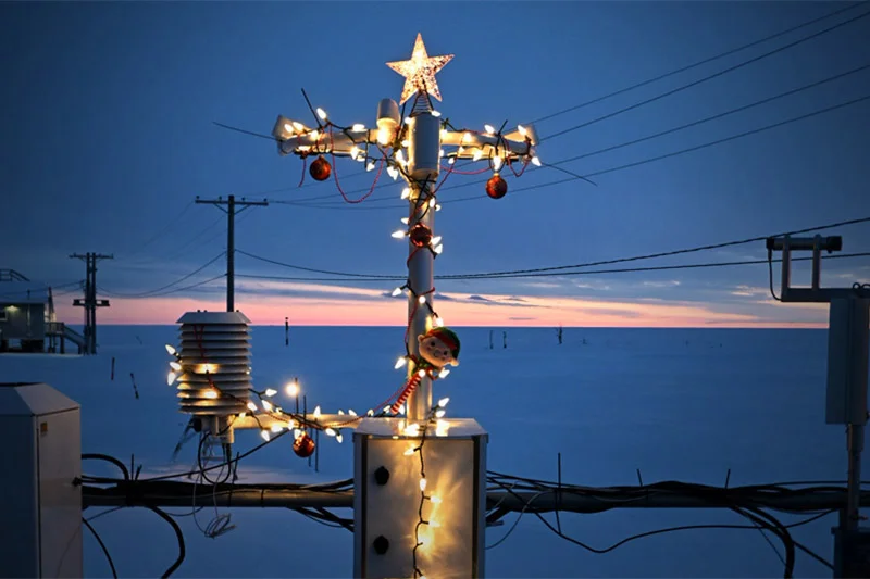 A utility pole and weather station are adorned with bright Christmas lights, a star, red ornaments, and an elf in snow at twilight.