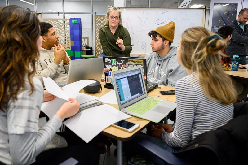 A female instructor in a green shirt gestures while speaking to four students with laptops and papers at a table in a bright classroom.