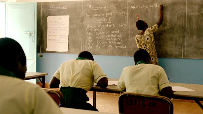 A teacher in a patterned dress writes a mathematics problem about determining profit margin on a classroom blackboard. Several students in cream and green uniforms sit at desks, facing the lesson.
