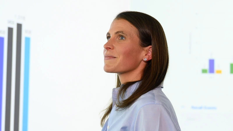 Brown-haired woman in a light blue collared shirt looking intently at colorful bar charts projected on a white wall. She has a subtle smile.