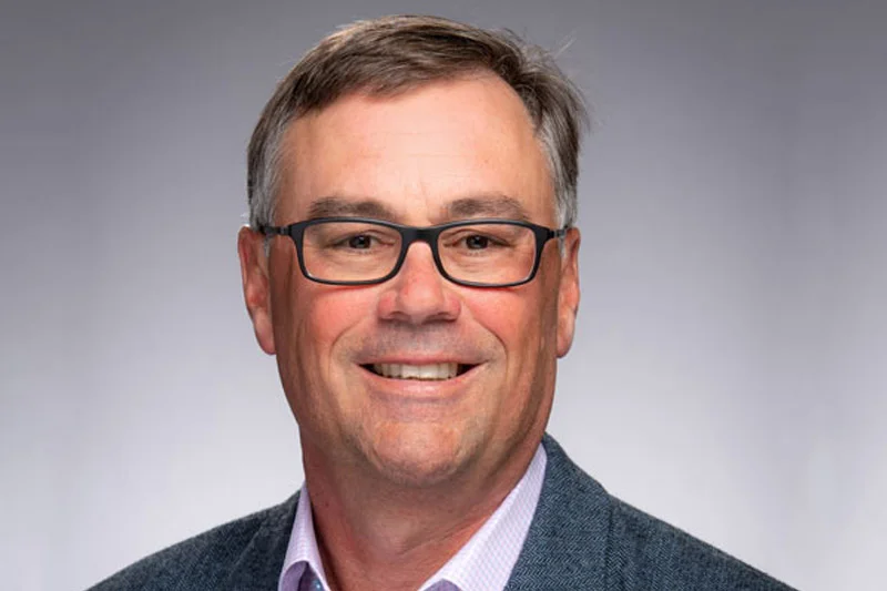 A smiling man with graying hair wearing black-framed glasses, a light purple shirt, and a dark gray tweed jacket.