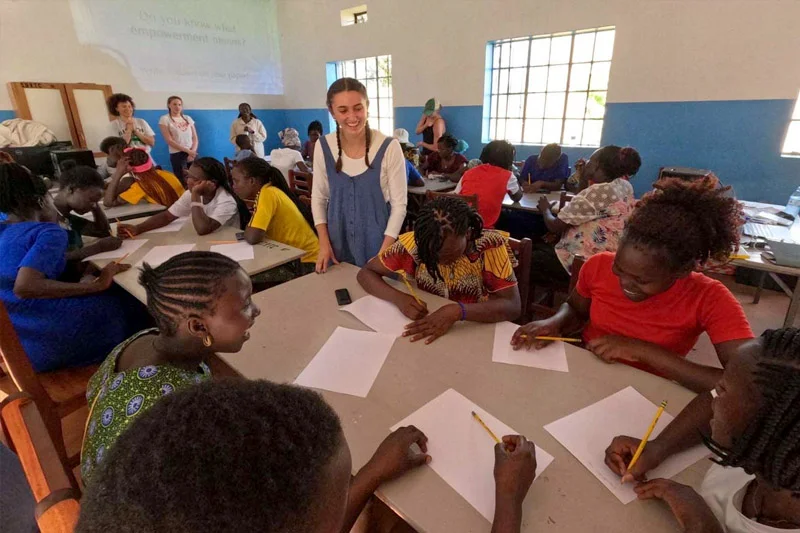 Smiling facilitator in blue overalls oversees women and girls writing about empowerment in a bright classroom.