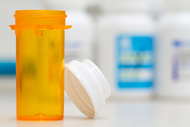 An empty orange prescription pill bottle with its white cap resting beside it sits on a counter in front of blurred medicine containers.