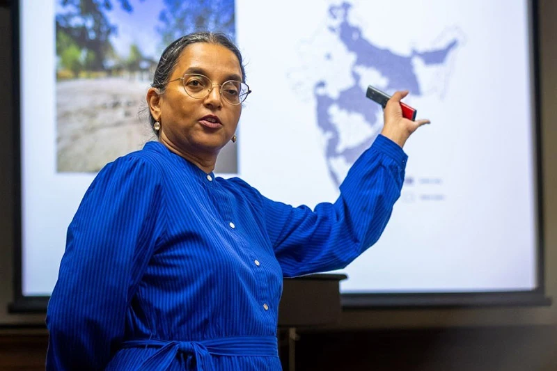 A woman in a blue dress and glasses gestures toward a projector screen showing a map of India and a landscape photo during a presentation. 