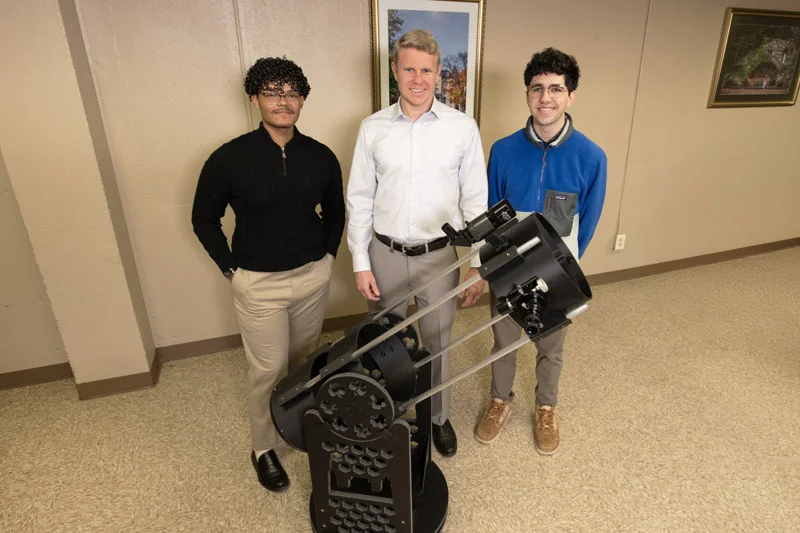 Three smiling men, one in a white shirt and one in a blue fleece, stand with a large black telescope.