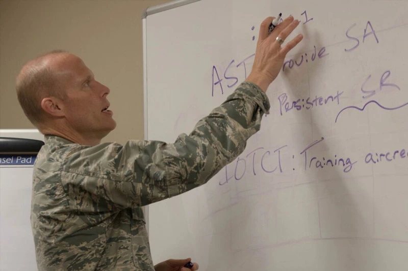 A man in military camouflage uniform writes notes on a white board with a black marker.