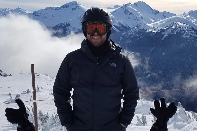 A man in a dark ski jacket, helmet, and goggles stands on a snowy mountain with peaks and clouds behind him.