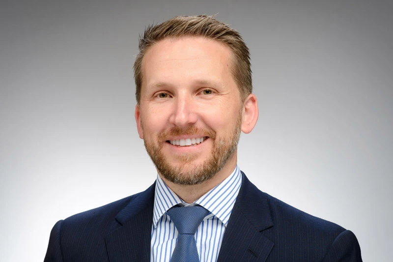 Headshot of a smiling man with a beard and light brown hair, wearing a navy pinstripe suit and a blue striped shirt and tie.