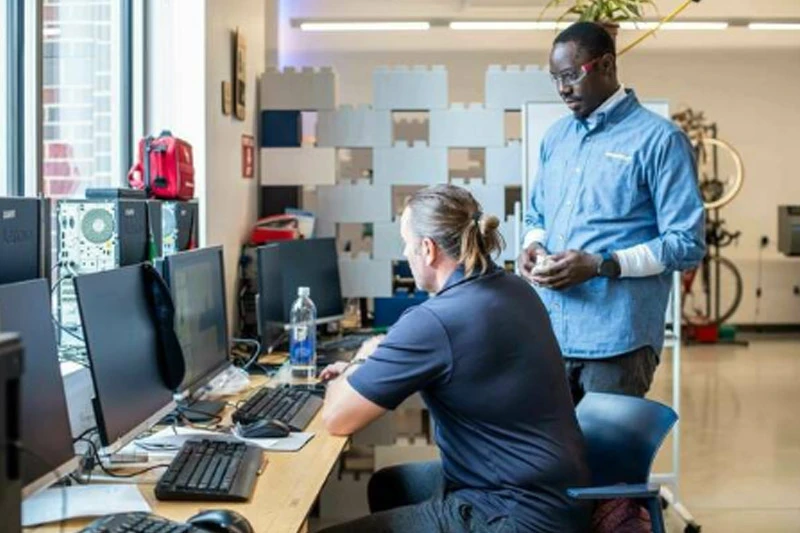 Two men work in a modern office, one seated at a multi-monitor computer and the other standing, looking over his shoulder.