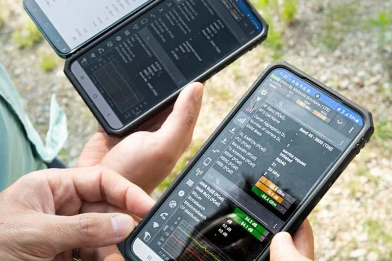 Hands hold two black smartphones outdoors displaying radio frequency signal strength and network data on dark screens. 