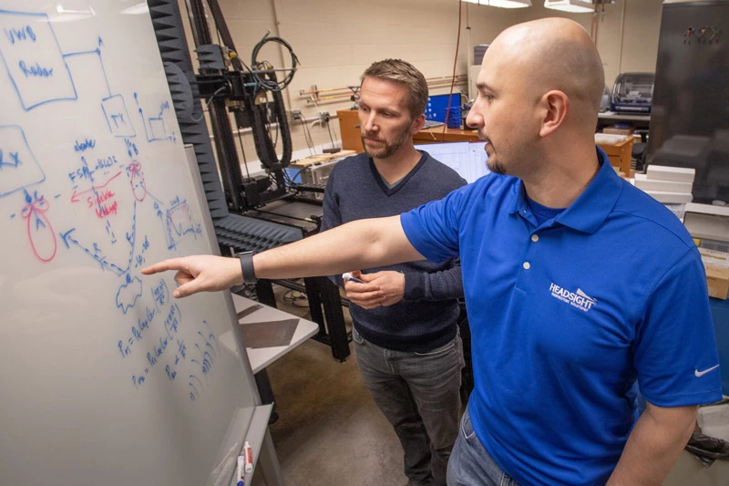 Two men, one pointing, discuss handwritten equations and diagrams on a white board in a laboratory setting.