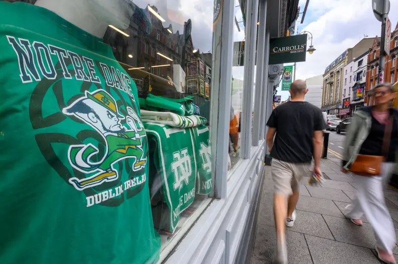A vibrant green Notre Dame t-shirt is displayed in a store window, with blurred pedestrians outside.