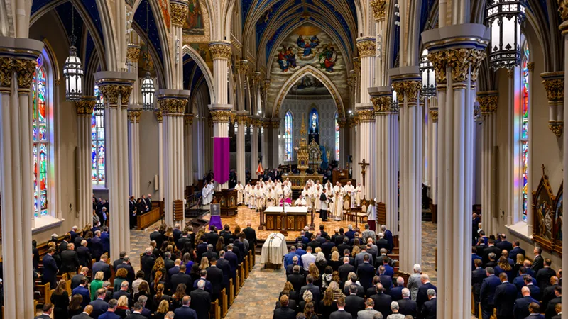 A packed funeral Mass at the Basilica of the Sacred Heart at the University of Notre Dame, with a white casket before the altar, priests in white vestments, stained glass windows, and the ornate blue and gold vaulted ceiling above.